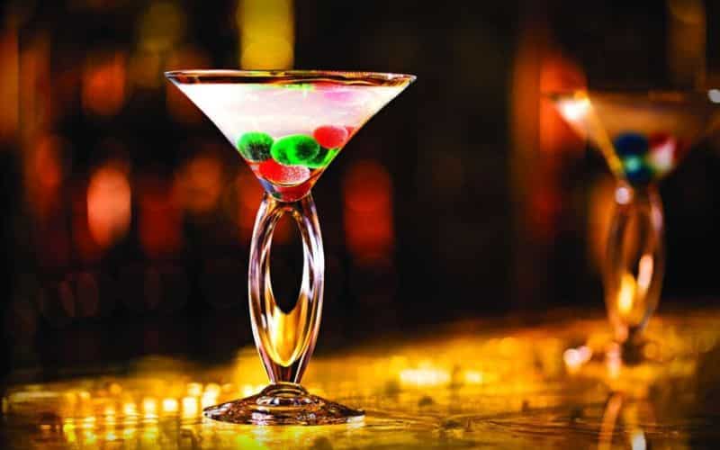 Two elegant martini glasses with a unique twisted stem design, the forefront glass filled with a clear liquid and colorful spherical garnishes, against a warm, bokeh-lit bar backdrop.