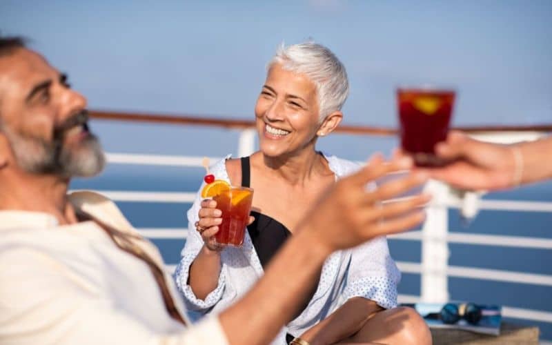 A smiling woman with short silver hair, joyfully toasting with a colorful cocktail on the deck of a ship, sharing a relaxed moment with companions against a clear blue sky. She is casually dressed, indicative of a leisurely cruise atmosphere.