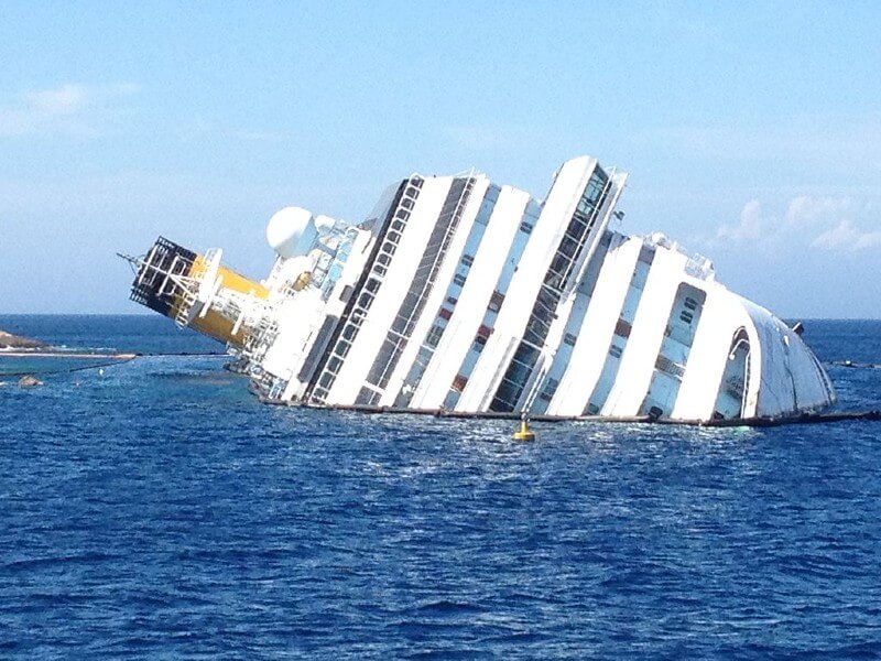 The Costa Concordia cruise ship lies capsized off the coast of Isola del Giglio, Italy, in bright daylight, displaying the stark aftermath of its 2012 maritime disaster.