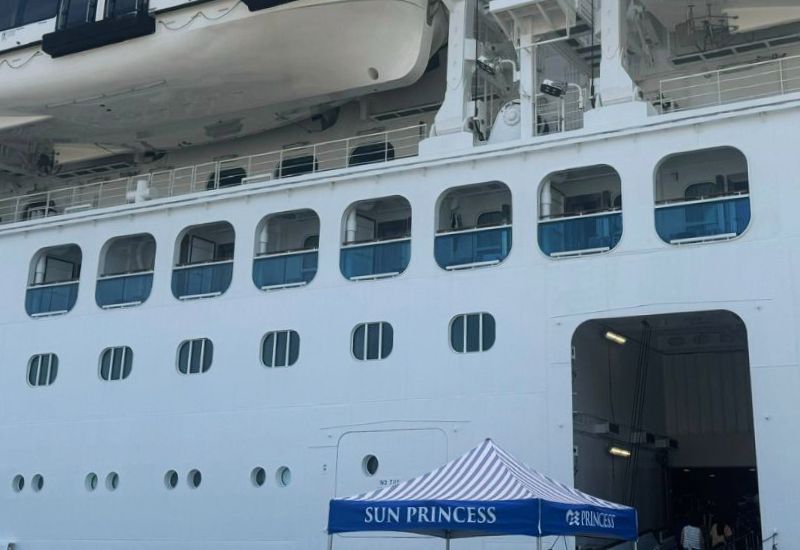 The image shows the exterior view of the Cove Balcony Staterooms on Princess Cruises' Sun Princess. These staterooms are located on a lower deck of the ship, identifiable by their distinctive covered balconies that are recessed into the ship's hull. The balconies provide a more sheltered outdoor space, offering privacy and protection from the elements while still allowing guests to enjoy ocean views. Above these staterooms, lifeboats are visible, indicating that these cabins are situated near the lifeboat deck. A "Sun Princess" canopy is set up near the gangway entrance, likely marking the boarding area for passengers.