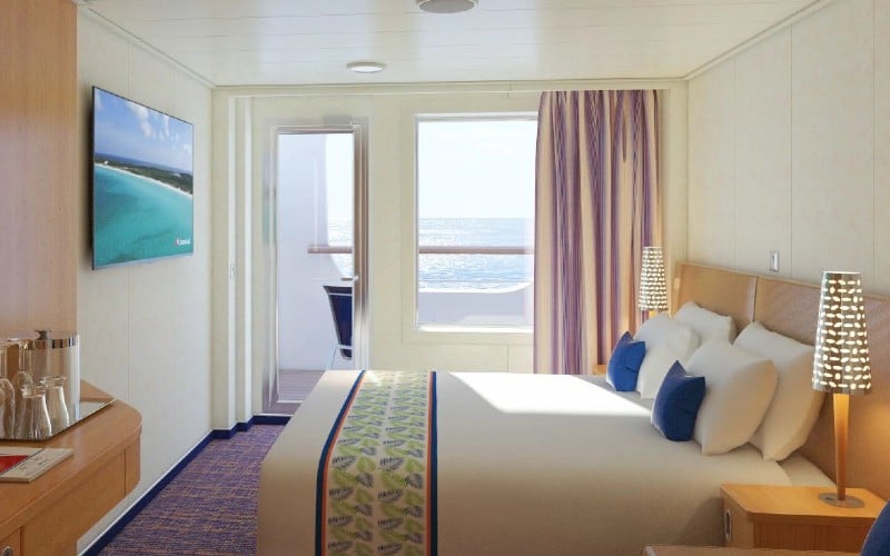 Cove Balcony stateroom on Carnival Horizon with a cozy bed, tropical-themed bedding, elegant bedside lamps, and a balcony door open to sea views, inviting a relaxing cruise experience.