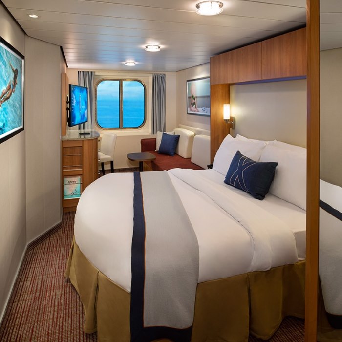 A snug three-person stateroom on a Celebrity cruise ship, featuring a large bed with a gold-and-navy accent, a porthole with a sea view, and a compact seating area, designed for efficient use of space.