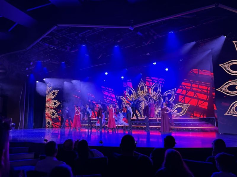 A vibrant live performance on the MSC Euribia stage, featuring a cast of dancers and singers in colorful costumes with intricate peacock feather designs in the backdrop. The stage is bathed in dramatic blue and purple lighting, enhancing the theatrical ambiance as the audience watches.