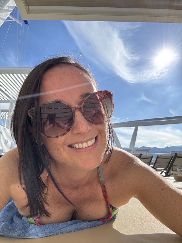 Cruise Mummy smiling with sunglasses reclining on a sun lounger on the deck of the Costa Smeralda cruise ship, with a clear blue sky and wispy clouds in the background.