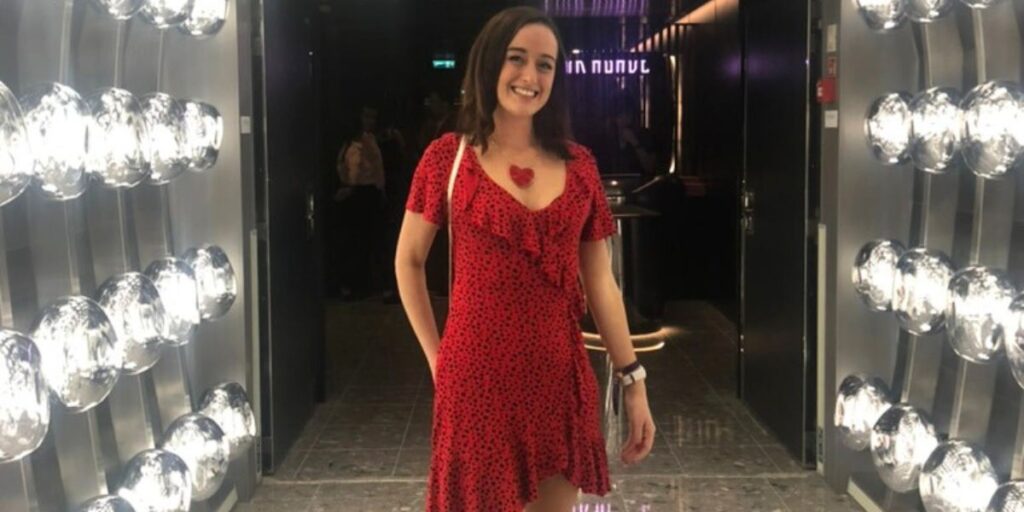 Cruise Mummy in a red polka dot dress with a heart-shaped pendant smiles while standing in a stylishly lit hallway on Virgin Voyages' Scarlet Lady. The modern decor features reflective surfaces and bright lights, creating a chic and vibrant atmosphere.