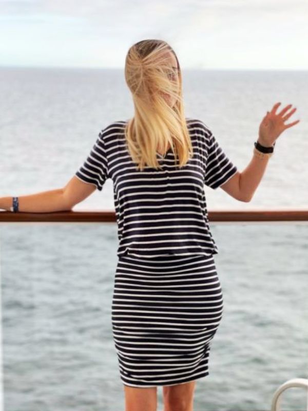 A woman in a striped dress standing on the balcony of the MSC Virtuosa, with her hair blowing in the wind and the ocean in the background.