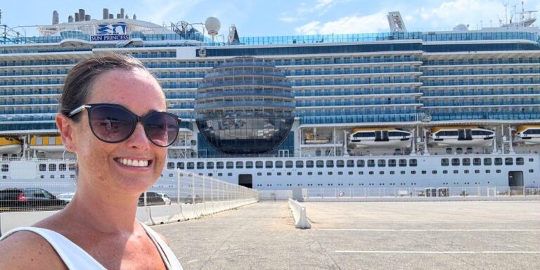 Cruise Mummy wearing sunglasses smiles at the camera while standing in front of the Sun Princess cruise ship. The massive ship is docked, showcasing its multiple decks, lifeboats, and the distinct spherical structure at its center. The bright day and clear sky add to the vibrant and exciting atmosphere as she prepares for her cruise adventure.