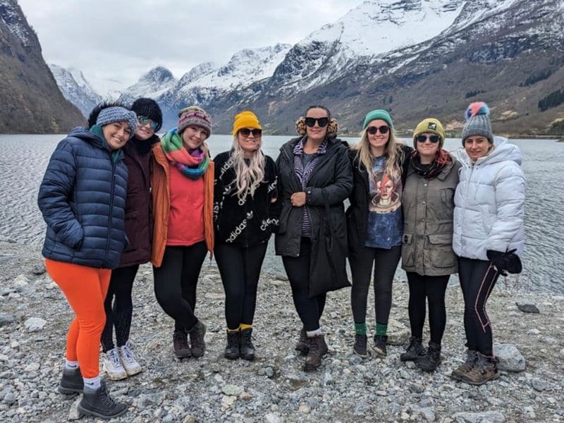 A group of cheerful cruise guests bundled up in warm attire pose together during a scenic shore excursion, with the majestic backdrop of a mountainous landscape and a serene lake, capturing a moment of camaraderie and adventure.