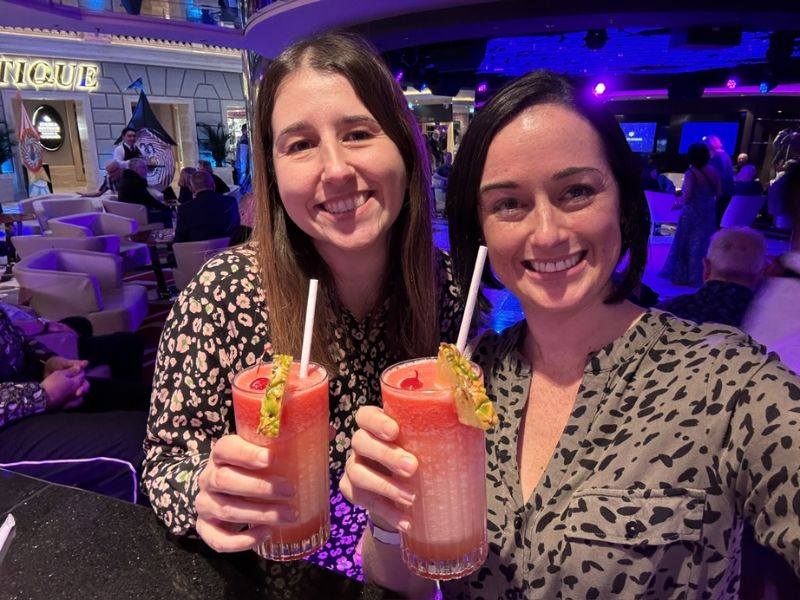 Two women smiling and toasting with vibrant pink cocktails adorned with fruit garnishes at a lively cruise ship bar. The atmosphere is festive with soft lighting and a blurred background of guests enjoying the entertainment.