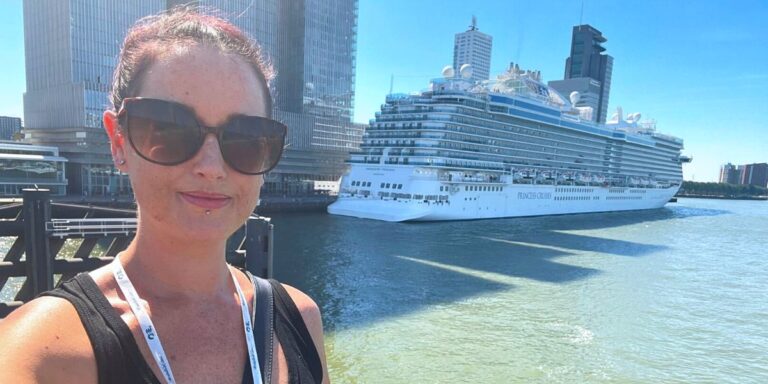 Cruise Mummy with sunglasses and a lanyard takes a selfie in front of a large Princess Cruises ship docked at a port, with tall buildings in the background under a clear blue sky.