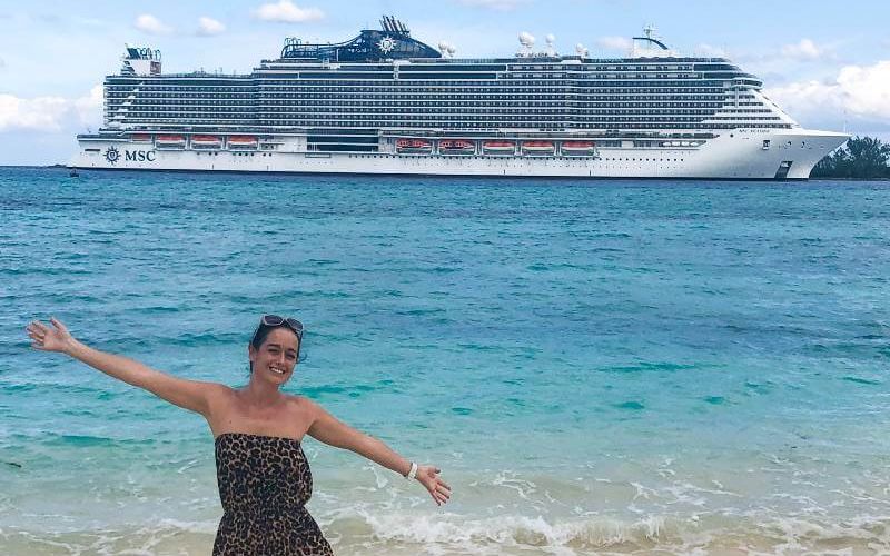 Cruise Mummy in a leopard print dress with sunglasses on her head, extends her arms joyfully on a sandy beach with the MSC cruise ship in the background, floating on calm turquoise waters under a partly cloudy sky.