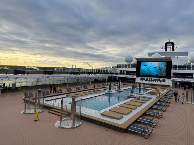 Early evening on MSC Euribia pool deck, featuring loungers, a tranquil pool with fountains, and a large outdoor movie screen displaying underwater imagery. The deck overlooks a port with industrial structures in the distance, under a soft sunset sky.