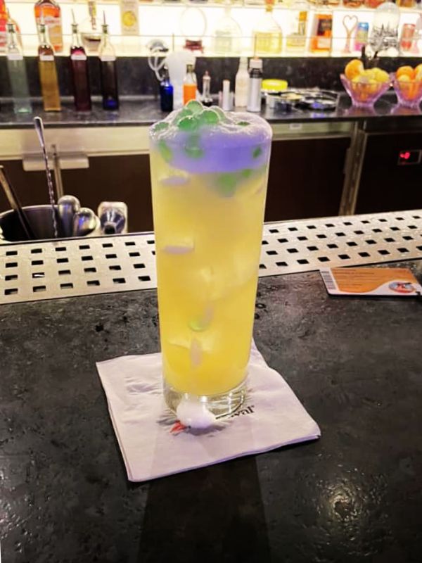 A Cucumber Sunrise cocktail on Carnival Cruise Line, featuring a tall glass filled with a yellow drink, ice, and green garnish. The drink is placed on a napkin with the Carnival logo, atop a bar counter with a backdrop of various bottles and bar utensils, creating a vibrant and inviting bar atmosphere.