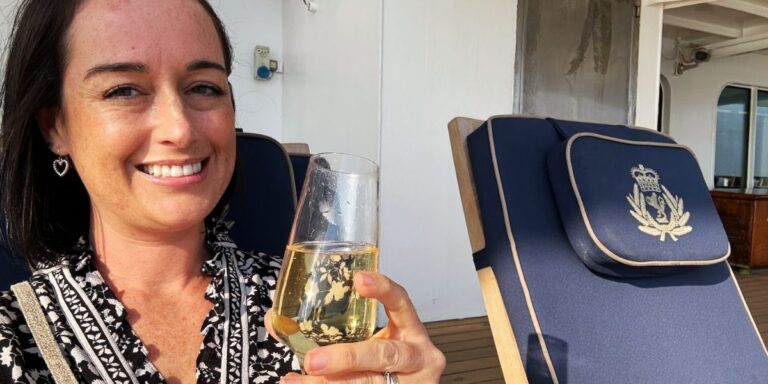 Cruise Mummy smiles while holding a glass of white wine on a Cunard cruise ship, seated on a deck chair with a navy cushion featuring a gold embroidered emblem.