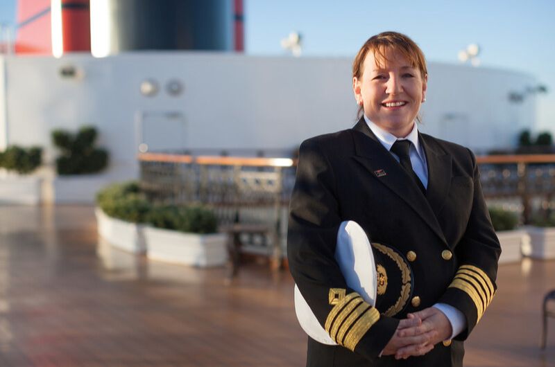 A Cunard cruise captain, dressed in a formal navy uniform with gold stripes on the sleeves, stands on the ship's deck holding a white captain's hat. The background features the ship's deck with potted plants and part of the ship's structure, under a clear blue sky, creating a professional and maritime ambiance.