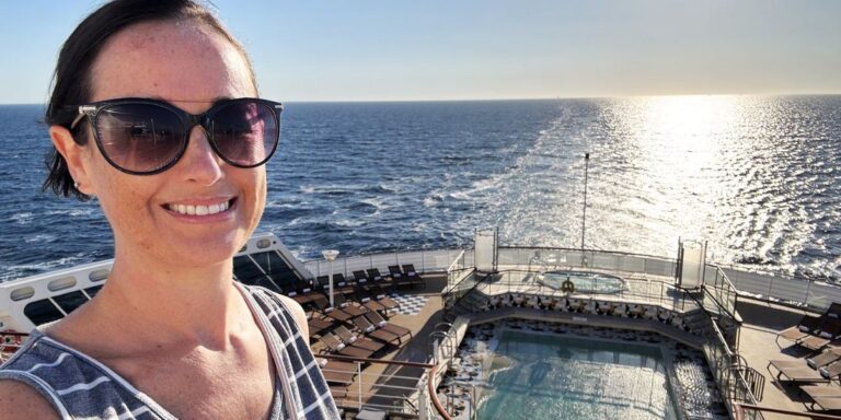 Cruise Mummy wearing sunglasses smiles at the camera while standing on the deck of the Cunard Queen Mary cruise ship, with the ocean and a setting sun in the background. The ship's deck features a pool and sun loungers.