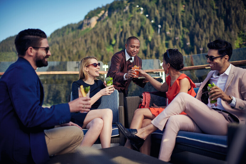 Cunard guests are enjoying drinks on the deck of a cruise ship in Alaska. The group, dressed in stylish attire, is seated on comfortable outdoor furniture with a scenic backdrop of lush green mountains. A waiter is serving cocktails, adding to the luxurious and relaxed atmosphere of the moment.