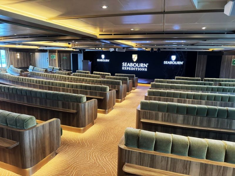 The Discovery Centre on the Seabourn Venture is designed as an elegant lecture hall where guests can attend educational talks and briefings. The room features tiered seating with plush green chairs, stylish wooden accents, and a modern wavy-patterned carpet. At the front, a large screen flanked by "Seabourn Expeditions" branding panels sets the stage for immersive learning experiences about the destinations the ship visits.