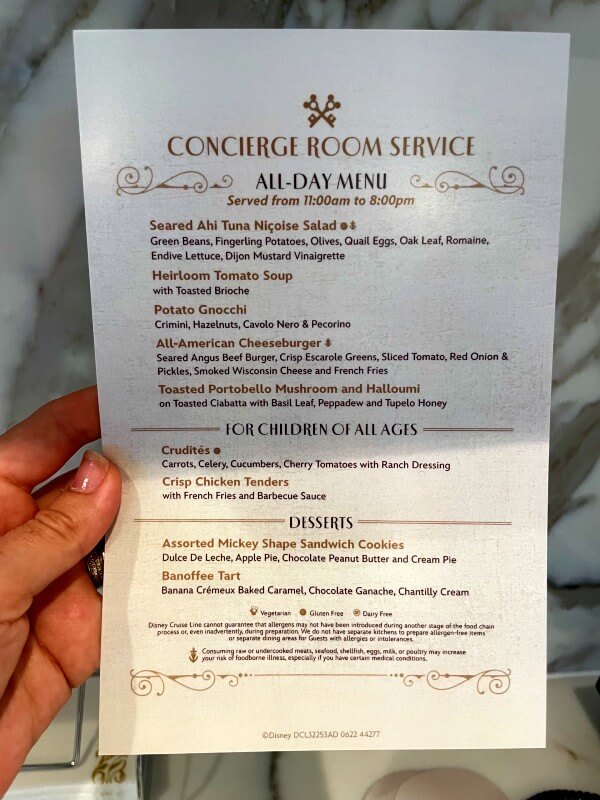 A detailed "Concierge Room Service All-Day Menu" from a Disney property, served from 11:00 am to 8:00 pm. The menu includes a variety of gourmet dishes such as Seared Ahi Tuna Niçoise, Heirloom Tomato Soup, and Potato Gnocchi with hazelnuts and Pecorino. It also features hearty options like an All-American Cheeseburger and Toasted Portobello Mushroom and Halloumi sandwich. For children, the menu offers simpler fare such as Crudités and Crisp Chicken Tenders. Desserts include Assorted Mickey Shape Sandwich Cookies and a Banana Crémeux Baked Caramel Tart. The menu is elegantly designed, featuring decorative fonts and borders, underscoring the upscale dining experience offered to guests.