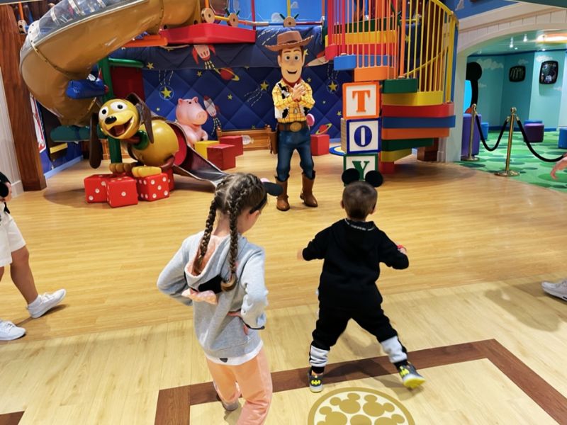 Children engaged in play at Disney Cruise Line's themed kids' facilities, with Woody from Toy Story greeting and interacting with young guests against a playful backdrop of colorful toy blocks and slides.