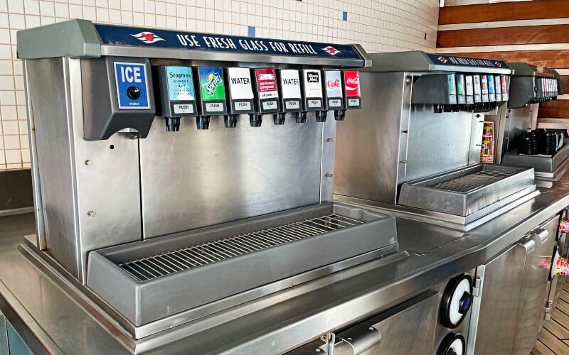 A row of drink dispensing machines on a Disney cruise ship, providing a variety of beverage options. These machines are labeled with popular soft drink brands such as Sprite, Coca-Cola, and Fanta, along with selections for ice and water. The area features a stainless steel counter and a reminder sign advising guests to "USE FRESH GLASS FOR REFILL", promoting hygiene and cleanliness. This setup is typical for cruise ships, offering guests convenient access to self-service beverages throughout their journey.