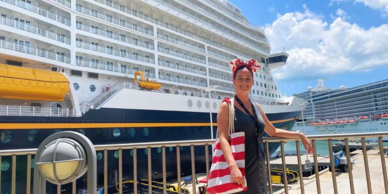 Cruise Mummy with a cheerful expression, dressed in a festive red and white striped outfit with a matching bow headband, poses at a port in front of a Disney Cruise ship. The ship's massive white structure, adorned with distinctive lifeboats in bright yellow, looms in the background, showcasing multiple decks and a playful, welcoming design typical of Disney Cruise Lines. The sunny weather and clear skies add to the vibrant, joyful atmosphere of the setting, perfect for a family adventure at sea.