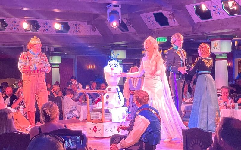 A vibrant performance of 'Frozen' characters in Arendelle: A Frozen Dining Adventure on the Disney Wish cruise ship, with guests enjoying the show featuring Anna, Elsa, Kristoff, and Olaf.