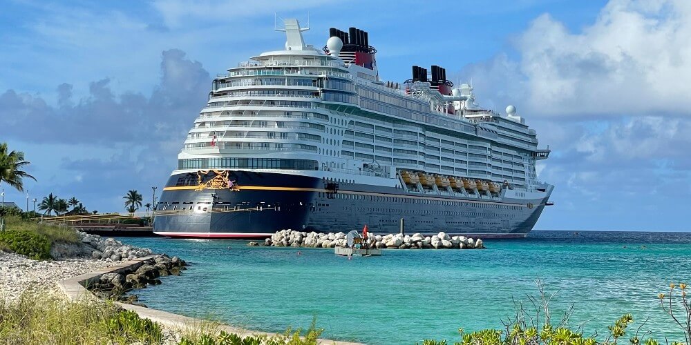 The Disney Wish cruise ship docked at Castaway Cay, with vibrant tropical foliage and the clear turquoise waters of the Bahamas visible.