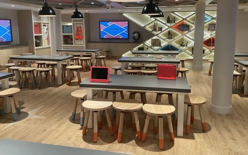 Spacious kids club room at Disney Cruise Line, featuring wooden tables and stools with orange accents, large screen displays with vibrant graphics, and illuminated shelving with decorative items in the background. The room is designed with a modern and playful aesthetic, perfect for children's activities and entertainment.