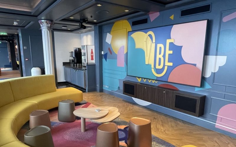 Vibrant teens club room on Disney Cruise Line, featuring a large curved yellow sofa, modern stools and tables, and a colorful wall mural with the word "Vibe" prominently displayed. The room also includes a beverage station and a large screen, creating a trendy and comfortable hangout space for teenagers.