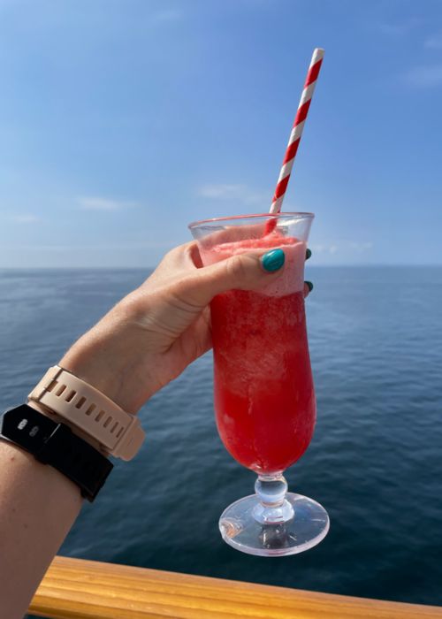 A hand with teal nails holding a frosted glass of red slush drink with a striped straw, set against a calm ocean backdrop, suggesting a relaxed vacation vibe.