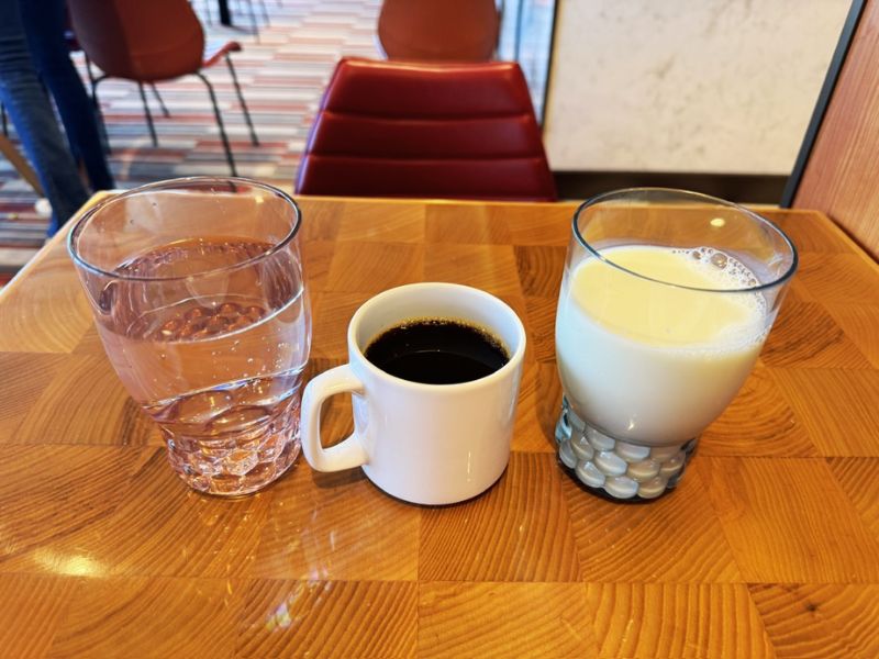 A refreshing trio of beverages on a wooden table, featuring a glass of water, a cup of black coffee, and a glass of almond milk, set against the interior of a dining area on the Costa Smeralda cruise ship.