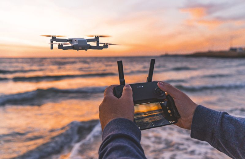 A person controls a drone with a remote, flying it over the beach at sunset. The drone captures the scenic view of the ocean waves and the colorful sky, creating a perfect moment for aerial photography.