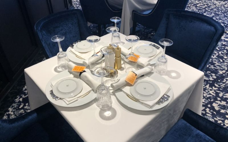 Formally set dining table on a NCL cruise ship, featuring white china, crystal stemware, and golden utensils, with place cards indicating early dining reservations, all atop a white tablecloth with a deep blue carpeted floor as backdrop.