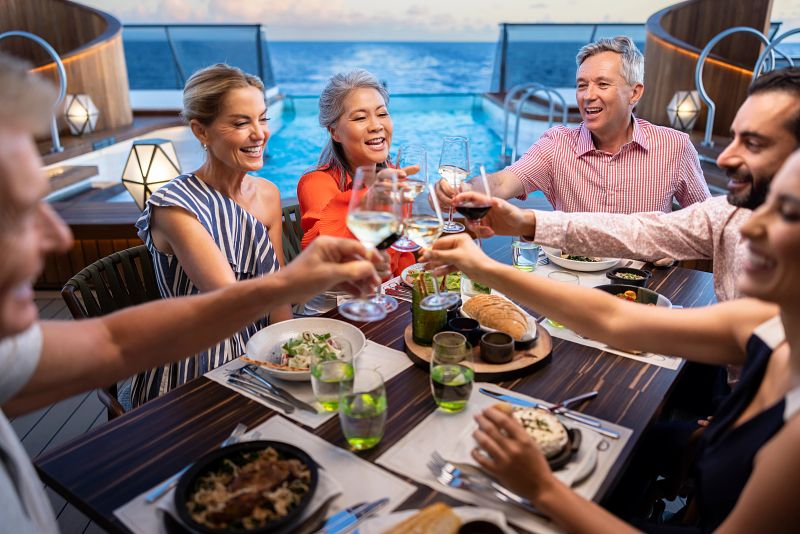 A group of cheerful guests enjoying a lively outdoor dinner at Earth & Ocean on a Seabourn cruise ship, toasting with glasses of wine amid a casual yet elegant setting with the ocean's endless horizon as the backdrop.