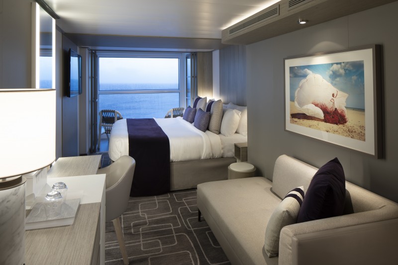 A luxurious Infinity Veranda stateroom on the Celebrity Edge cruise ship, featuring a plush bed with purple accents, a stylish seating area, and a modern desk. The room offers a stunning ocean view through the floor-to-ceiling window that can be opened to create a balcony experience, enhancing the spacious and elegant ambiance.