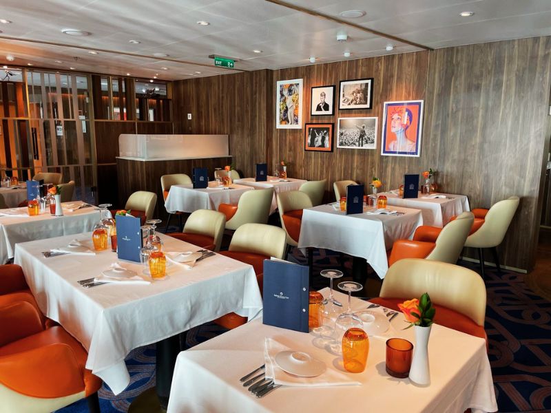 The Club Orange Dining Room on the Rotterdam, a Holland America Line cruise ship, featuring an inviting arrangement with two-toned chairs, set tables with orange water pitchers, and a selection of black-and-white and color art pieces on the walls.