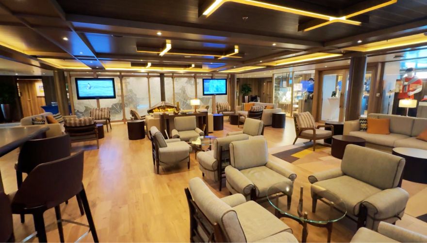 The Expedition Lounge on the Seabourn Venture is designed as a sophisticated and inviting space for relaxation and socializing. The lounge is outfitted with plush, light-colored armchairs and sofas grouped around sleek glass coffee tables, creating a comfortable environment for guests. Ambient lighting from elegant wooden panels and overhead geometric lights enhances the warmth of the space. Additionally, large screens displaying oceanic and map visuals provide an immersive backdrop, complementing the exploratory theme of the lounge.