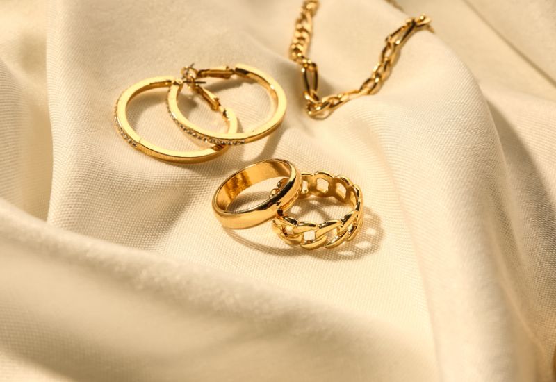 A collection of gold jewelry, including two rings, a bracelet, and a pair of hoop earrings, is elegantly displayed on a smooth, cream-colored fabric. The jewelry pieces shine under soft lighting, emphasizing their luxurious and expensive appearance.