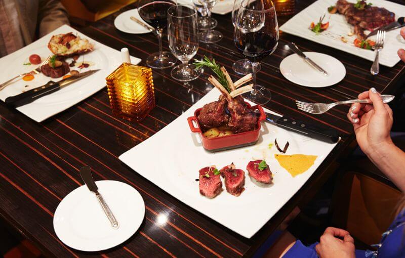 The image displays a fine dining setting at Fahrenheit 555 Steakhouse on a cruise ship. Plates with gourmet dishes, including perfectly cooked cuts of meat like lamb chops and steaks, are arranged on a dark, elegant table. The table is set with wine glasses, cutlery, and a small candle, creating a sophisticated and inviting dining atmosphere.