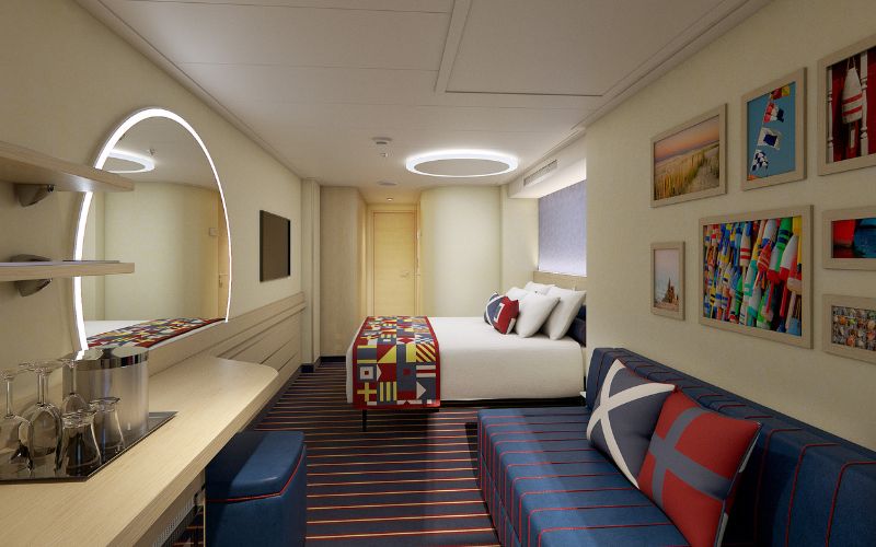 Interior view of a Family Harbor stateroom on a Carnival cruise ship, featuring a cozy bed with a nautical theme, a bench with vibrant patterns, a curved work desk with a mirror, and a wall adorned with colorful maritime-themed artwork.