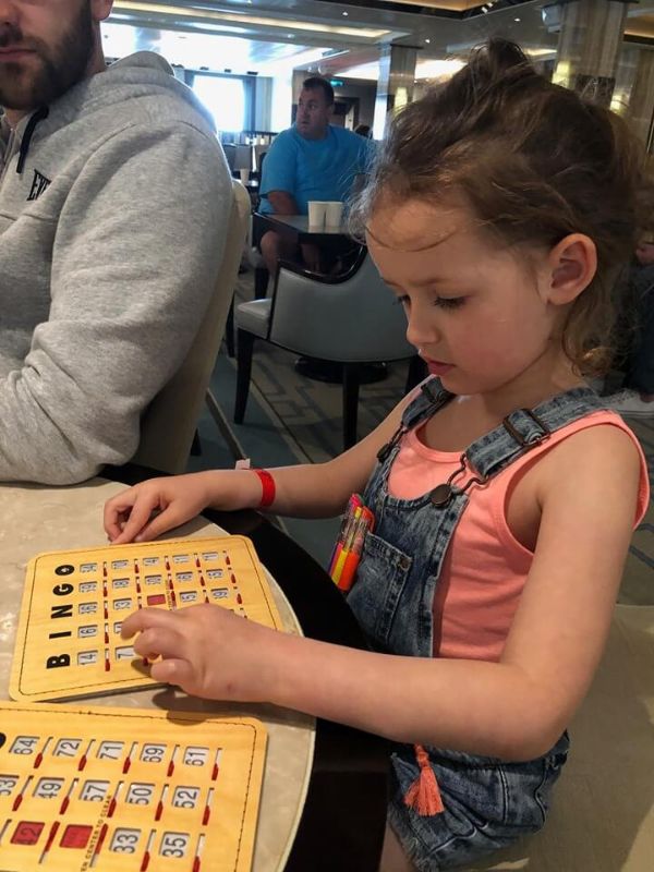 A young girl in a pink top and denim overalls is focused on playing bingo, marking her card with multiple colored markers, in a social setting with people in the background.