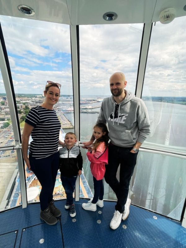 A family enjoying their vacation together, standing inside the glass-enclosed viewing area on a Royal Caribbean cruise ship with a panoramic view of the port and city in the background.