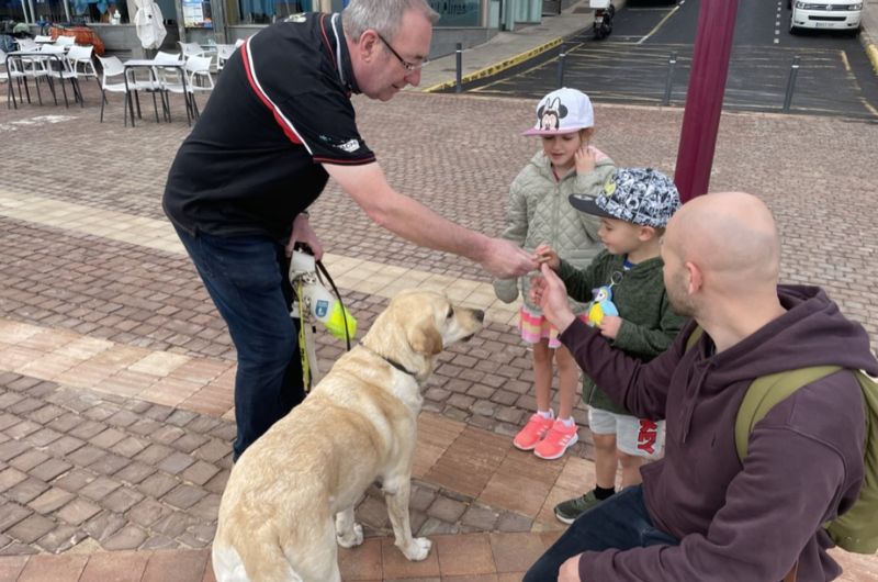A man in a black shirt introduces a light-colored Labrador Retriever guide dog to my children and husband seated nearby, on a paved area with chairs and tables in the background. The dog attentively receives a treat from the man's hand.