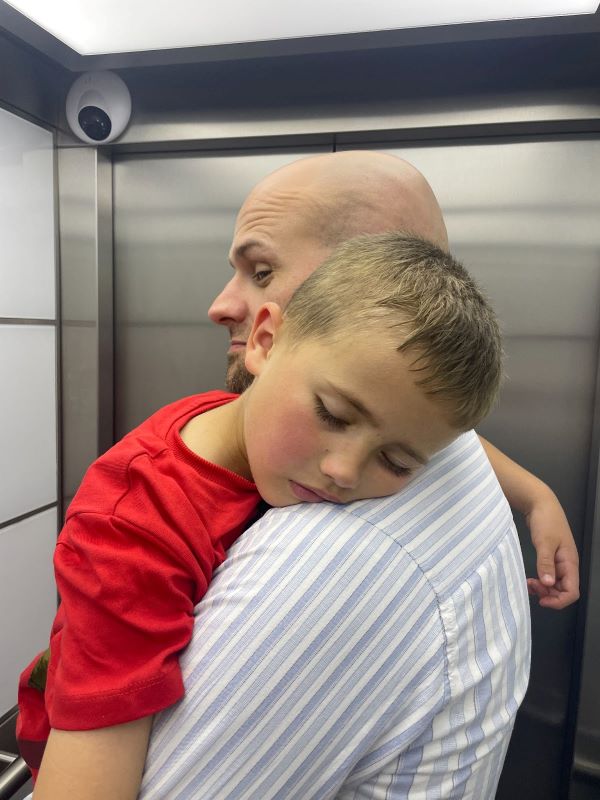 A tender moment as a father carries his tired son in a red shirt, both in the quiet space of a cruise ship elevator, after a day in the kids' club.