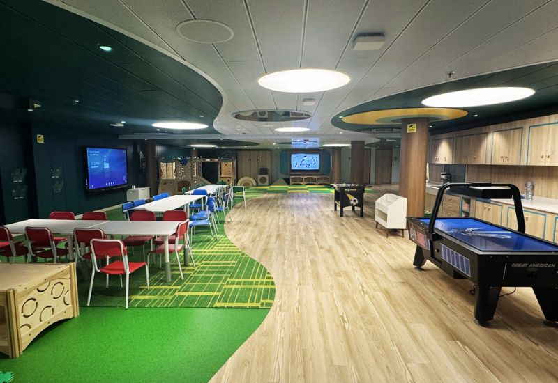 The image shows Firefly Park on Princess Cruises' Sun Princess, which appears to be a children's play area or activity center. The space is brightly lit and features a variety of activities, including long tables with red and blue chairs, an air hockey table, and a foosball table. There are also interactive screens on the walls, likely for gaming or educational activities. The floor is a mix of green carpet and wood-like vinyl, with playful, modern decor that creates a fun and engaging environment for kids. The area is spacious and well-organized, designed to keep young passengers entertained.