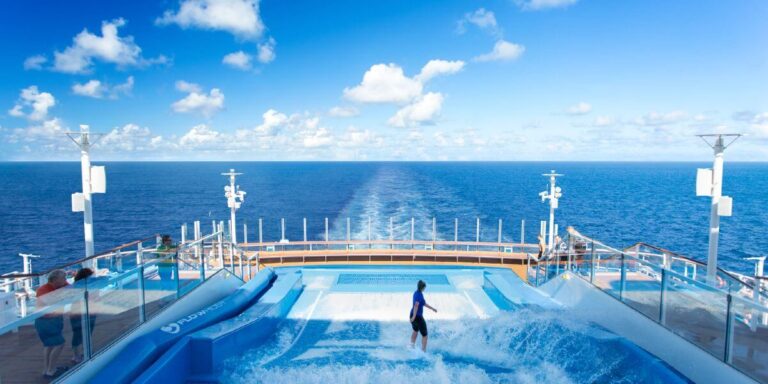 A woman is surfing on the FlowRider aboard Anthem of the Seas, enjoying the onboard wave simulator. She is balancing on the artificial wave while the ship sails through the open ocean, leaving a trail of white wake behind. The scene is set under a bright blue sky with scattered clouds, creating a thrilling and picturesque moment of adventure on the cruise ship.