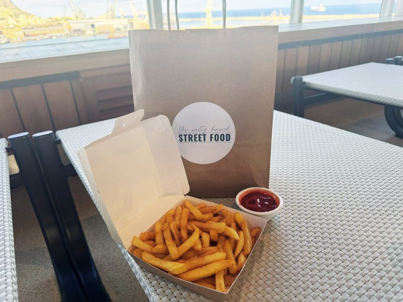 A casual dining setup at The Salty Beach Street Food cafe on Costa Smeralda, featuring a box of crispy golden fries with a side of ketchup, and the cafe's branded paper bag on a wicker table with a view of the harbor in the background.