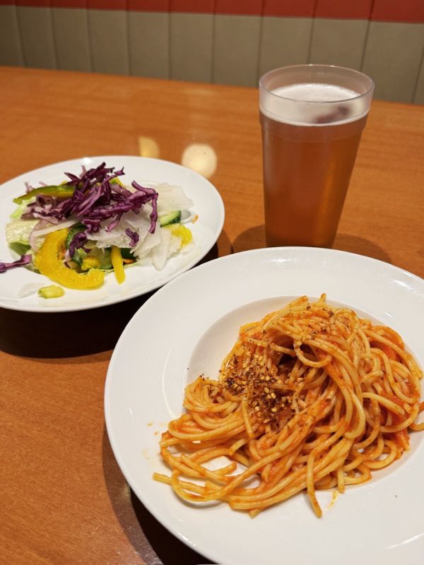 A plate of spaghetti with a rich tomato sauce and a sprinkle of herbs, next to a side salad with mixed greens and a glass of amber-colored beer, served on a table aboard the Costa Smeralda cruise ship.