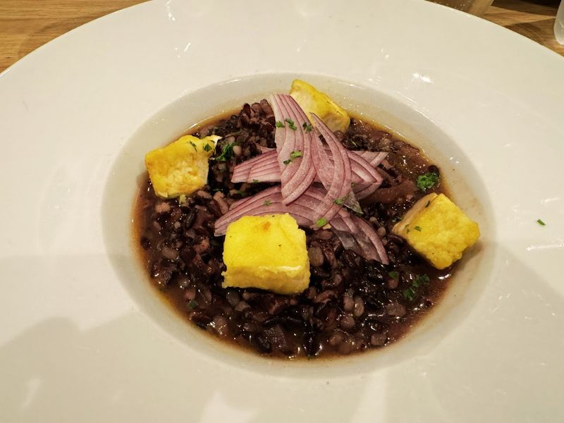 A gourmet dish of seasoned black lentils topped with crispy polenta cubes and sliced red onions, served on a white plate, highlighting the fine dining experience aboard the Costa Smeralda.